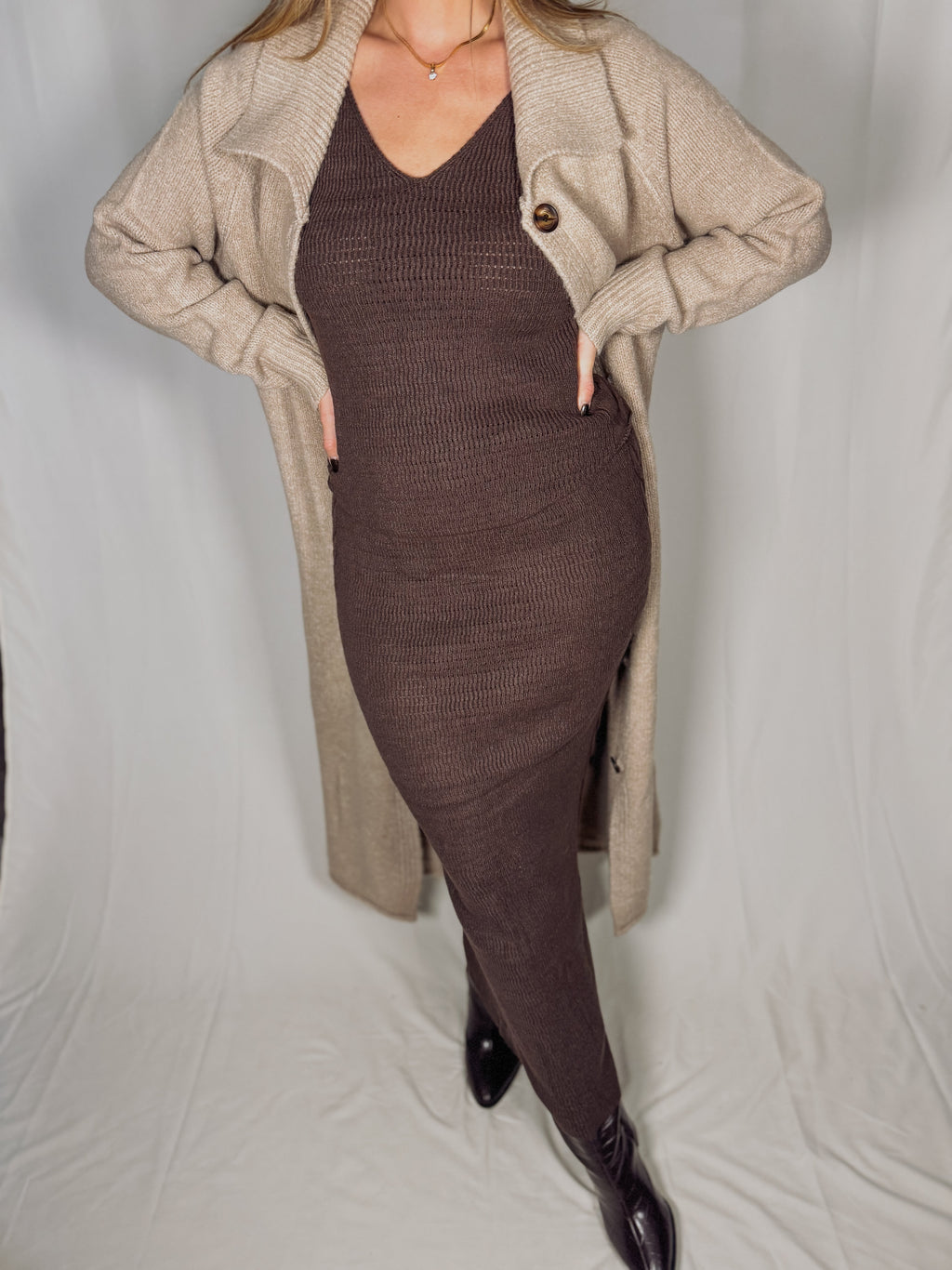 Person wearing a long beige coat over a brown tank and skirt set on a plain background