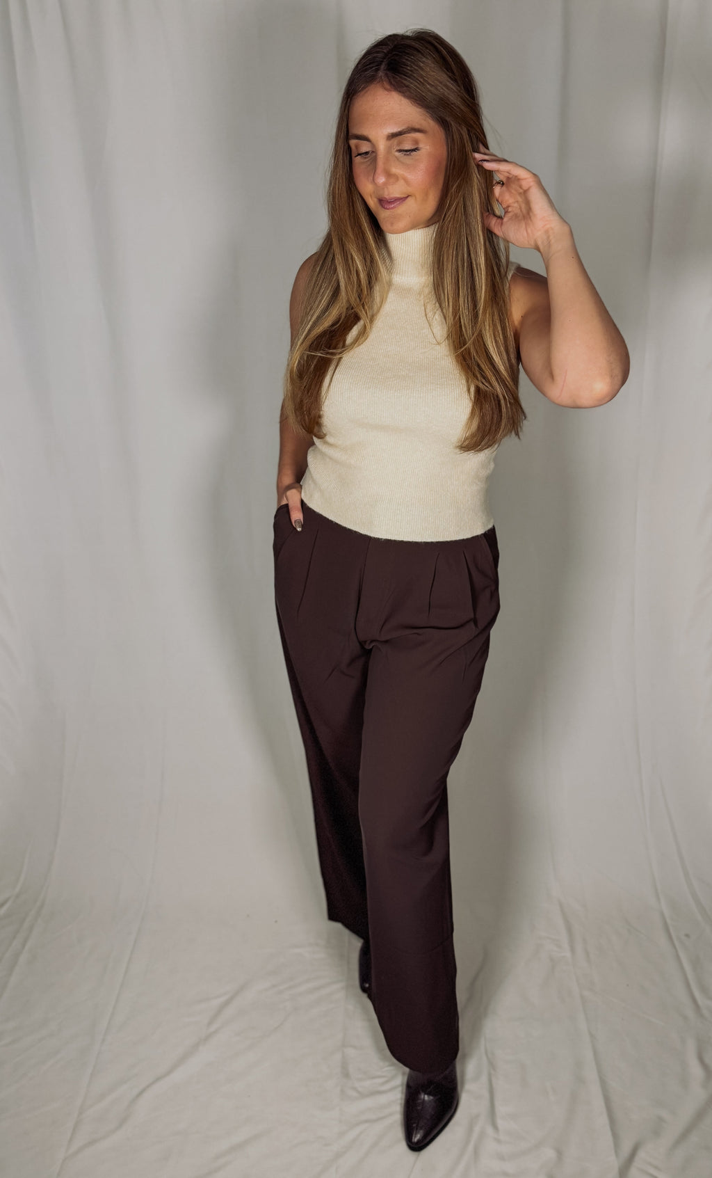 Woman wearing a cream top and dark brown pants against a plain background