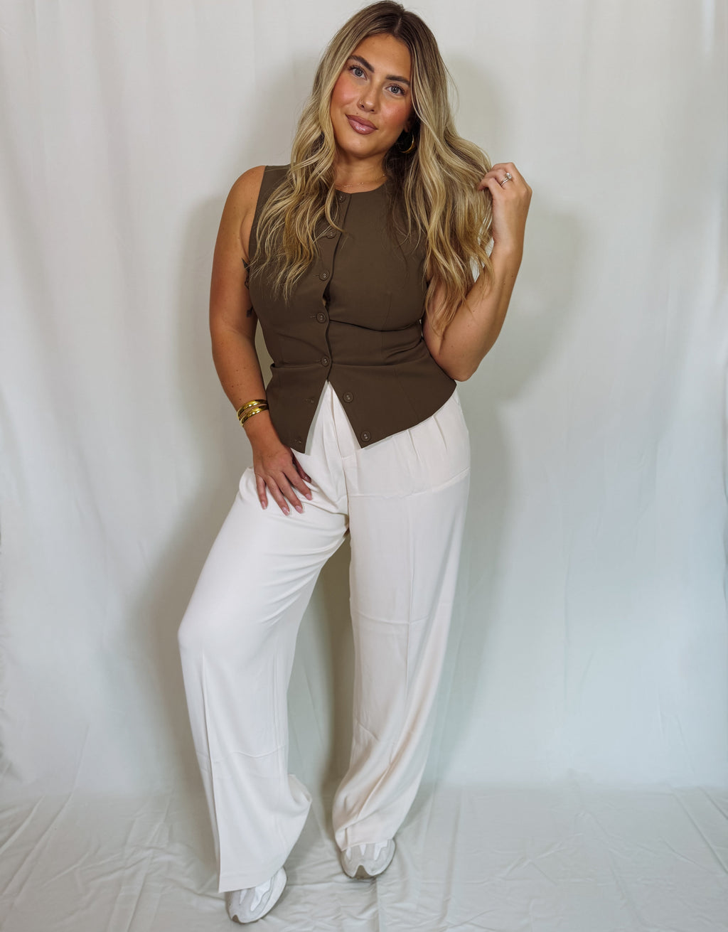 Woman wearing a brown sleeveless top and white pants against a plain background