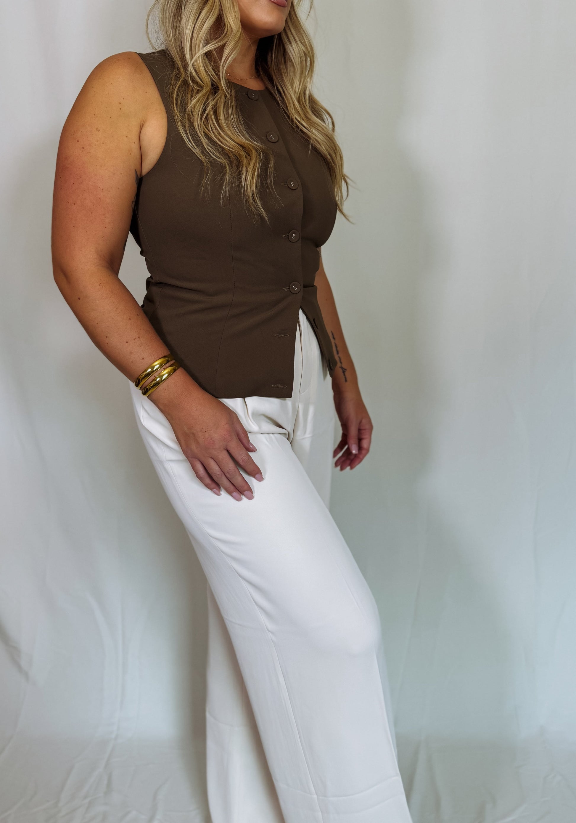 Woman wearing a brown sleeveless top and white pants against a plain background