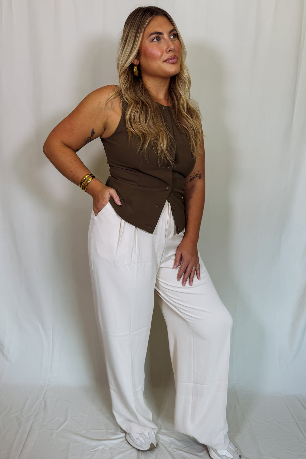 Woman wearing a brown sleeveless top and white pants against a plain background