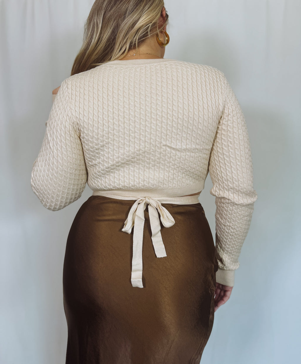 Person wearing a beige textured sweater and brown satin skirt against a plain background