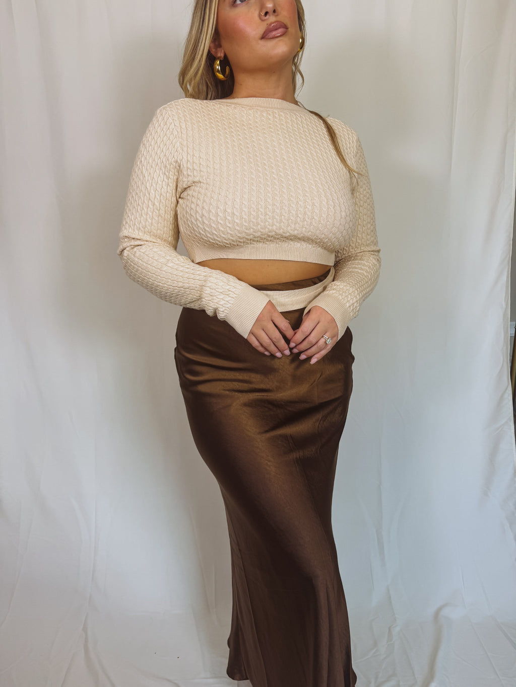 Woman wearing a beige ribbed sweater and brown satin skirt against a white background