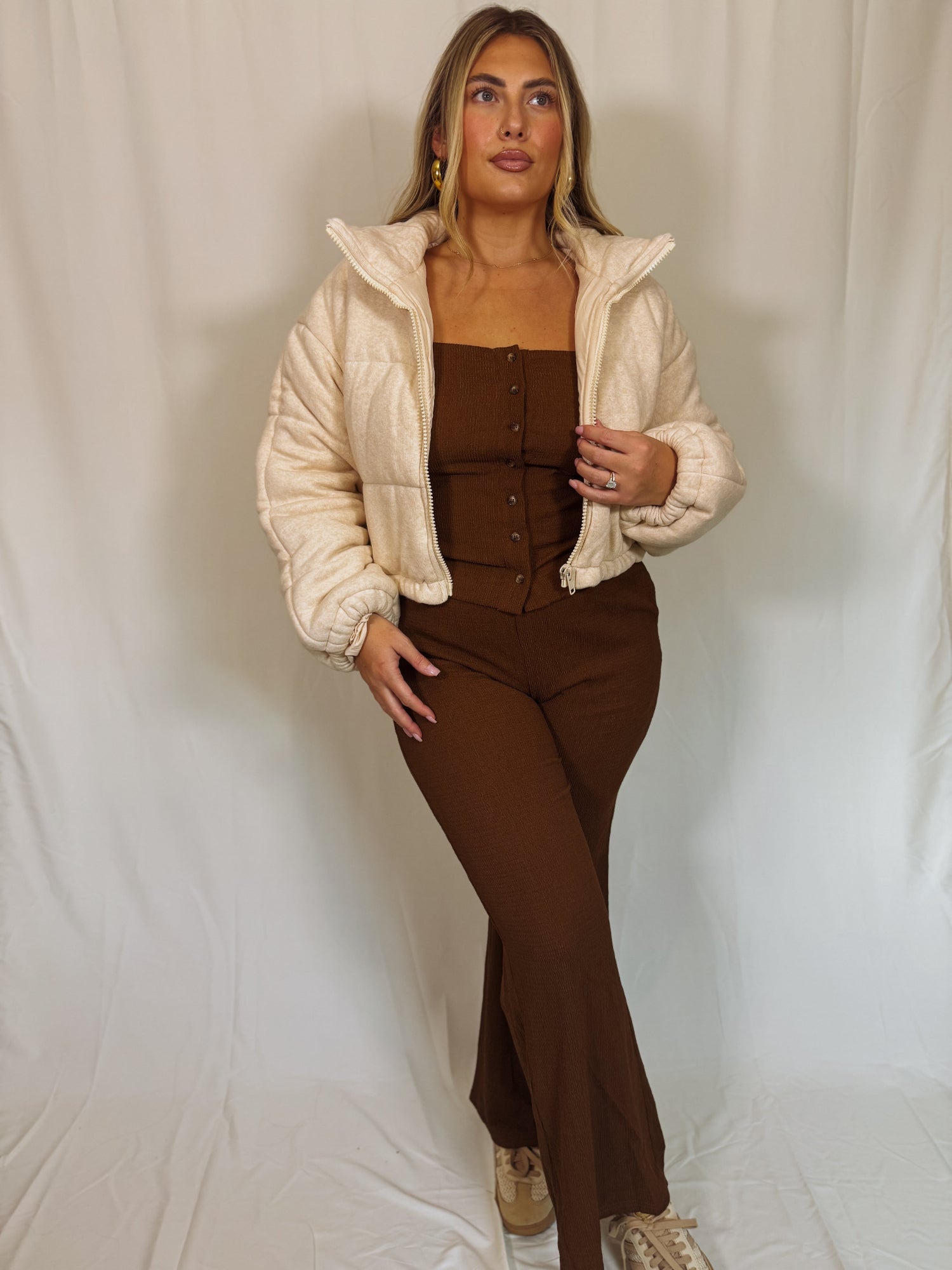 Woman wearing a beige puffer jacket over a brown outfit against a plain background