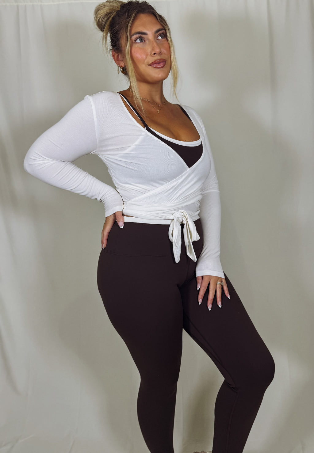 Woman wearing a white long-sleeve top and brown leggings against a plain background
