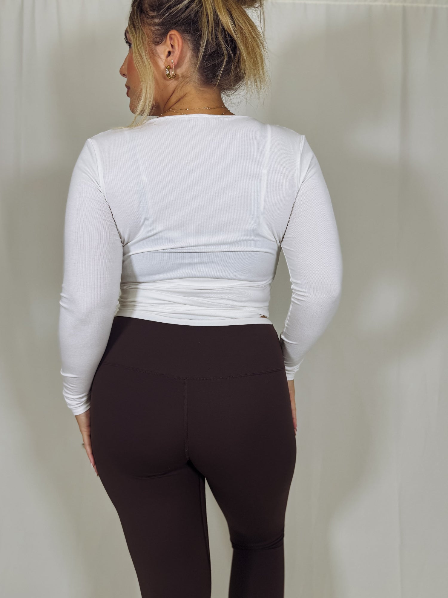 Person wearing a white long-sleeve top and brown leggings against a plain background