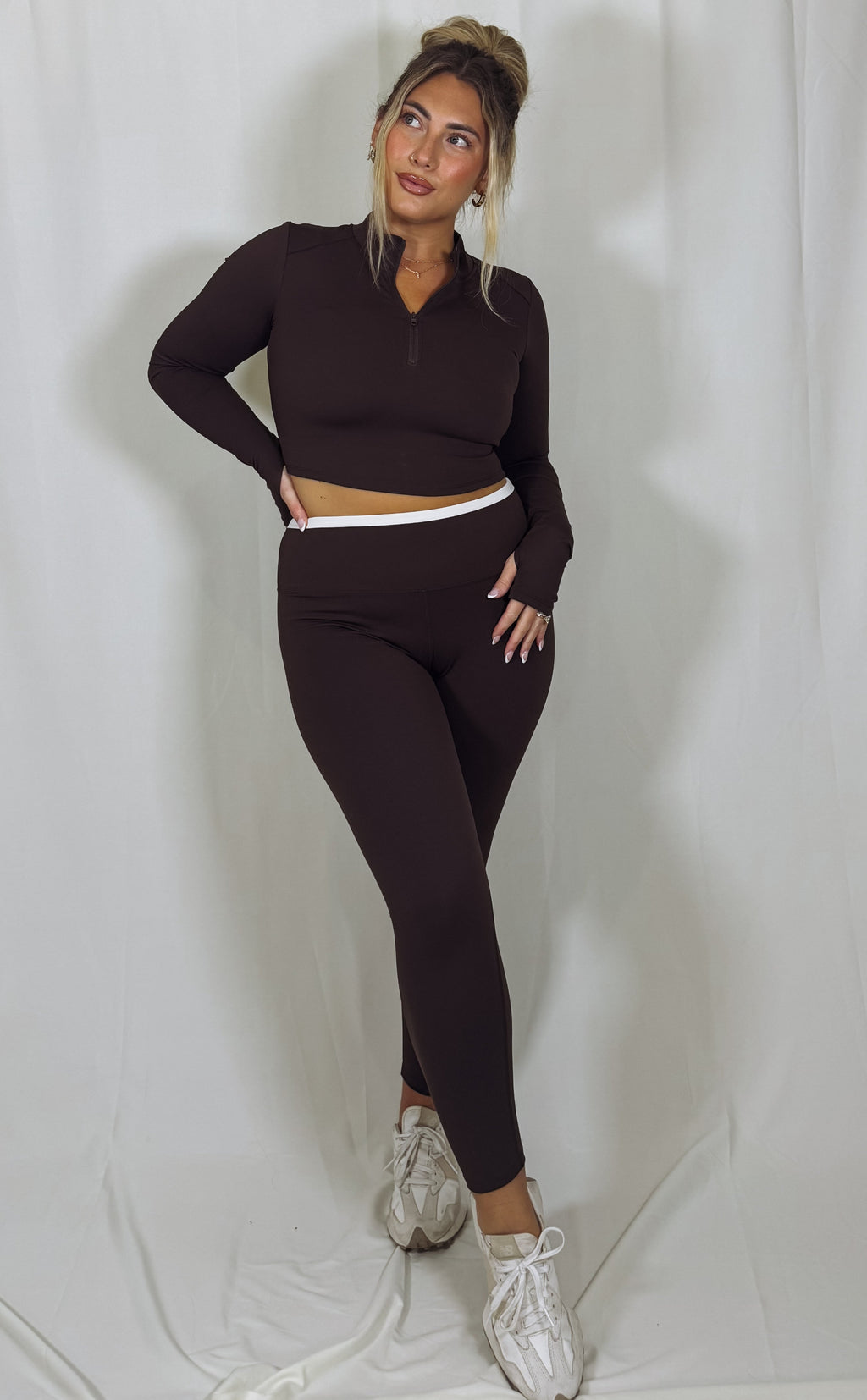 Woman wearing a dark brown long-sleeve top and leggings set with white sneakers on a light gray background