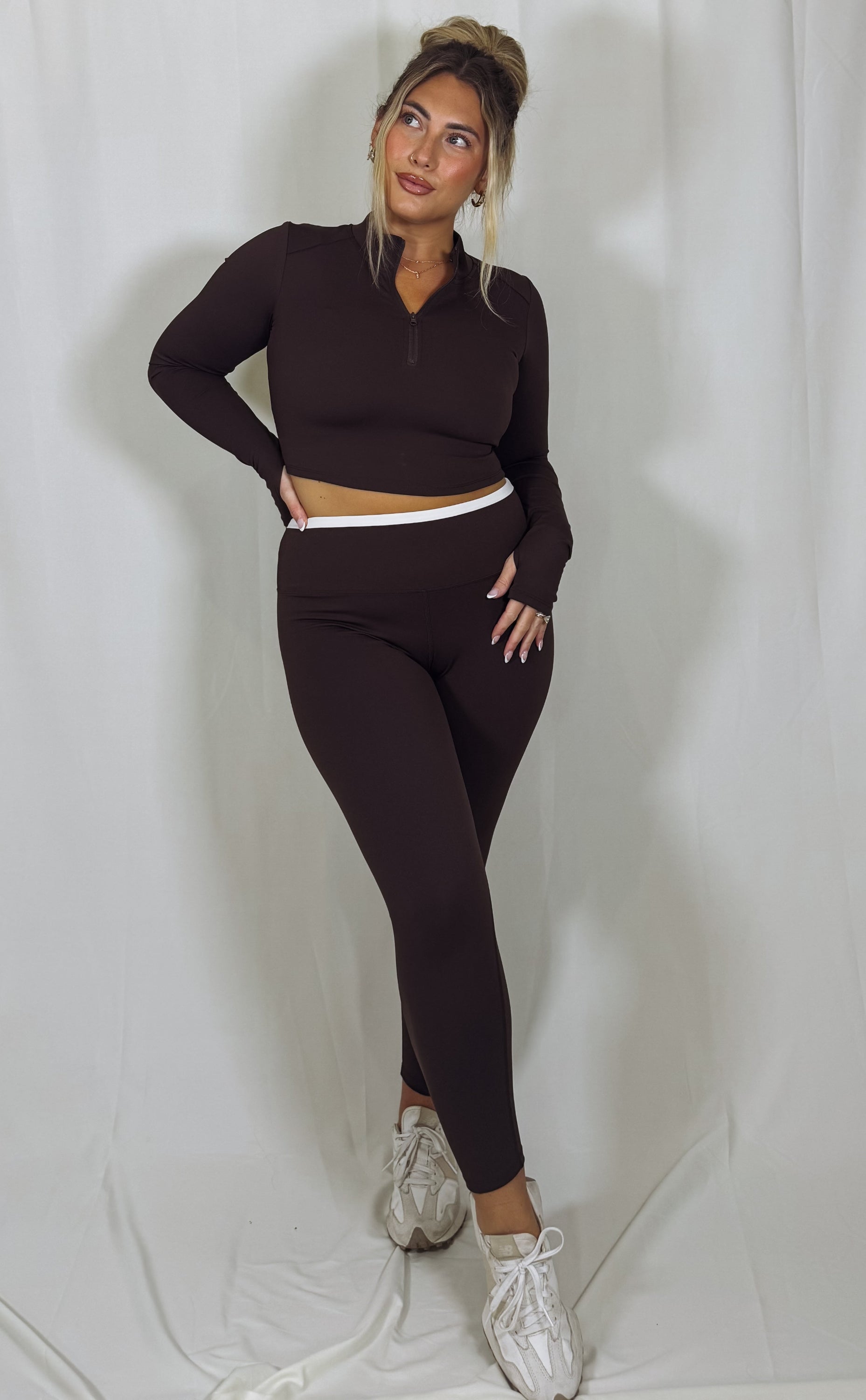 Woman wearing a dark brown long-sleeve top and leggings set with white sneakers on a light gray background