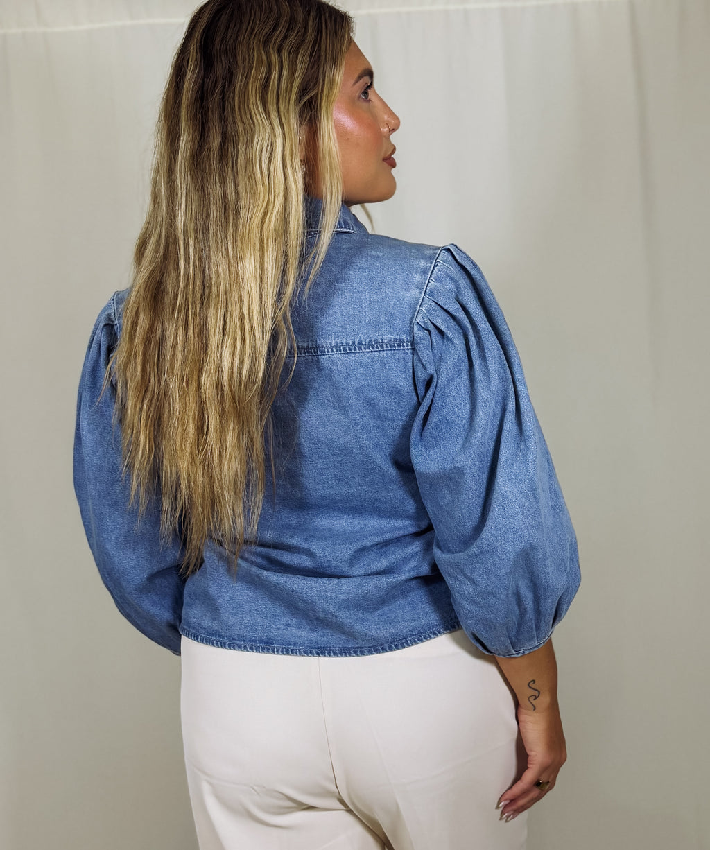 Person wearing a blue denim shirt with puffed sleeves against a beige wall.